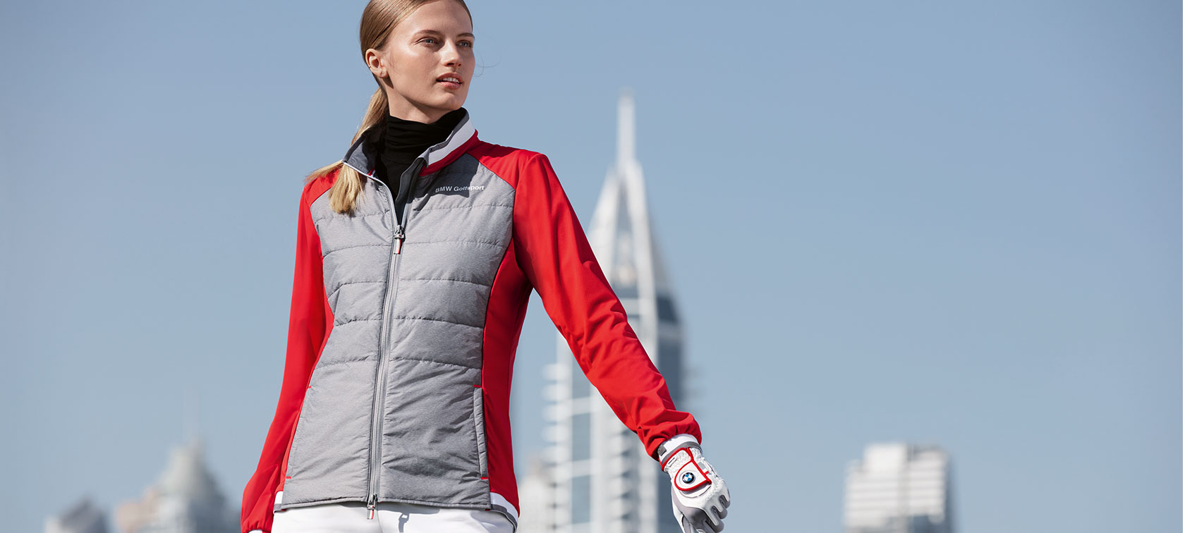 Dámská bunda BMW Golfsport. Mladá žena na obrázku s pozadím velkoměsta ukazuje produkty z kolekce BMW Golfsport.