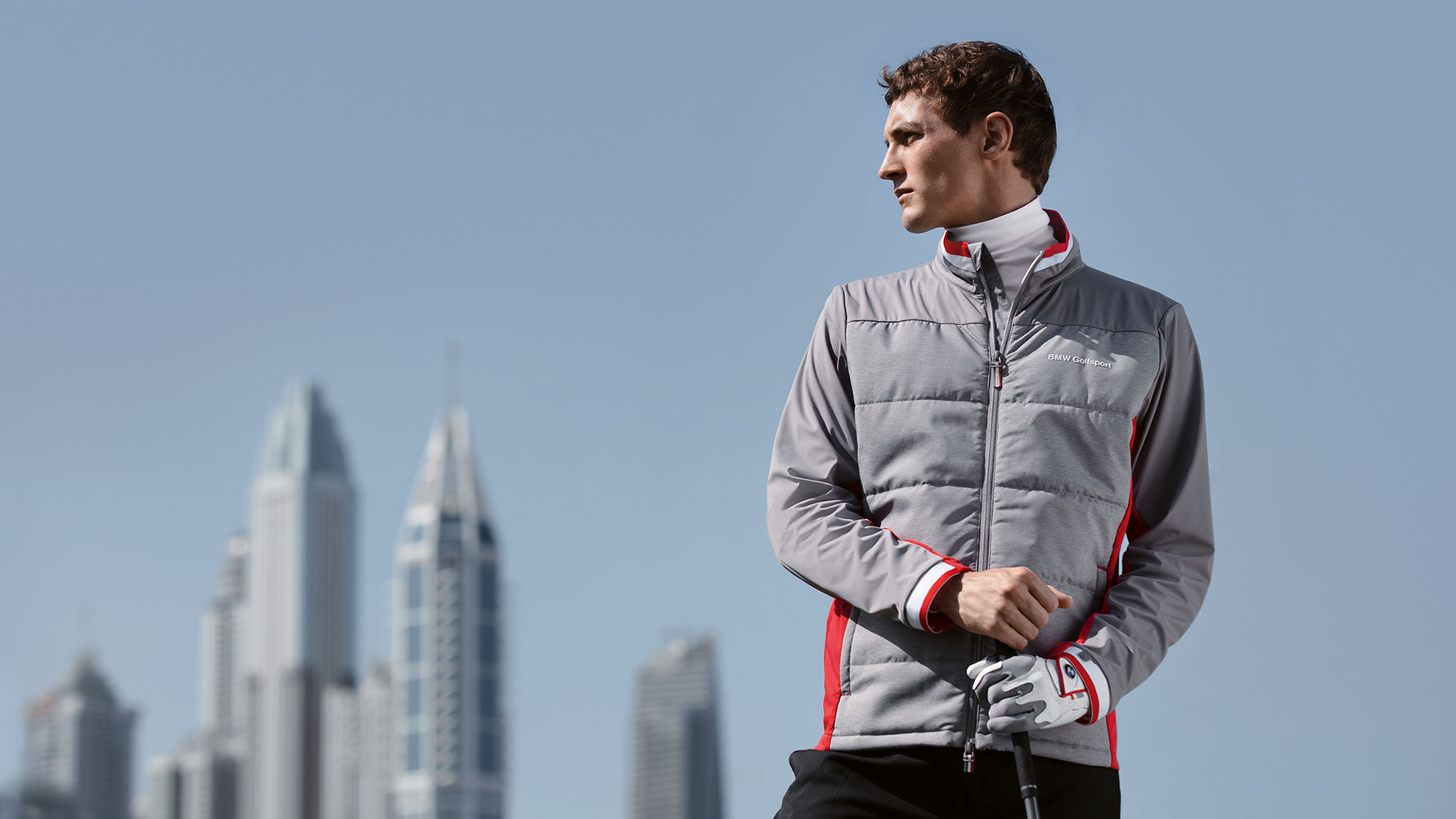 Muž předvádí produkty z kolekce BMW Golfsport Collection na pozadí velkého městského panoramatu. Muž předvádí produkty z kolekce BMW Golfsport Collection na pozadí velkého městského panoramatu.