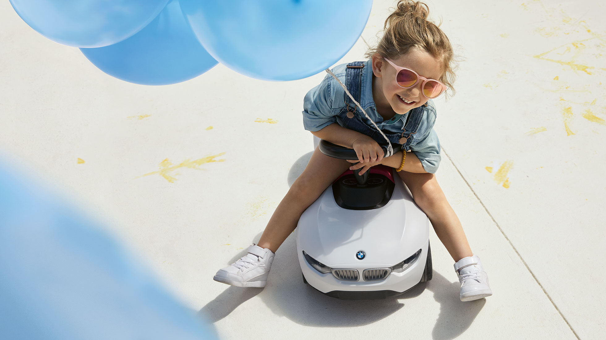 Kolekce BMW Kids. Dívka s růžovými brýlemi a modrým balonem radostně sedí na dětském odrážedle BMW Baby Racer.