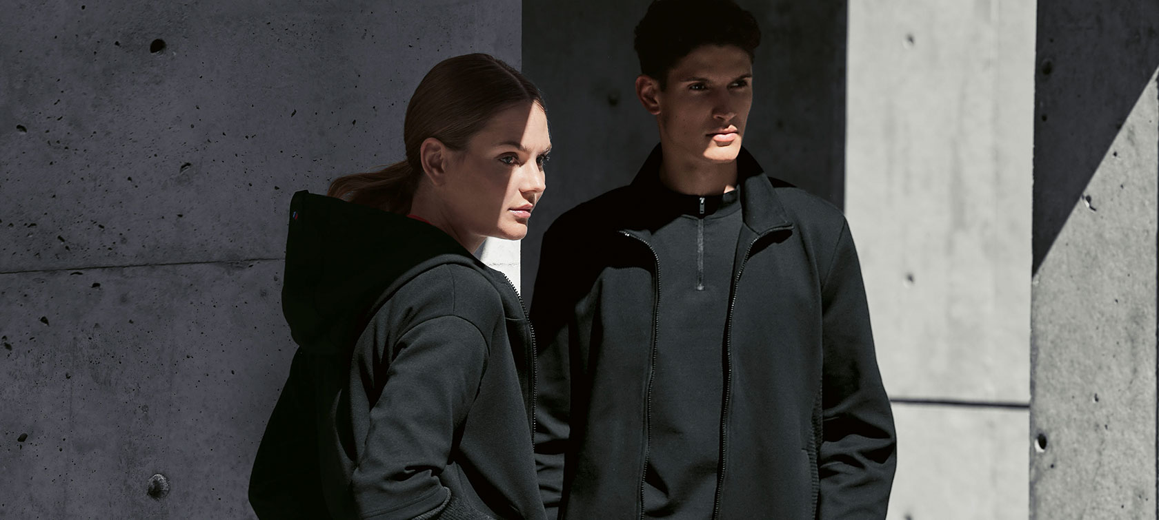 Dámská bunda BMW M Sweat Jacket a pánská bunda BMW M Sweat Jacket Pár stojí ve stínu. Žena předvádí dámskou bundu BMW M Sweat Jacket a muž má na sobě pánskou bundu BMW M Sweat Jacket.