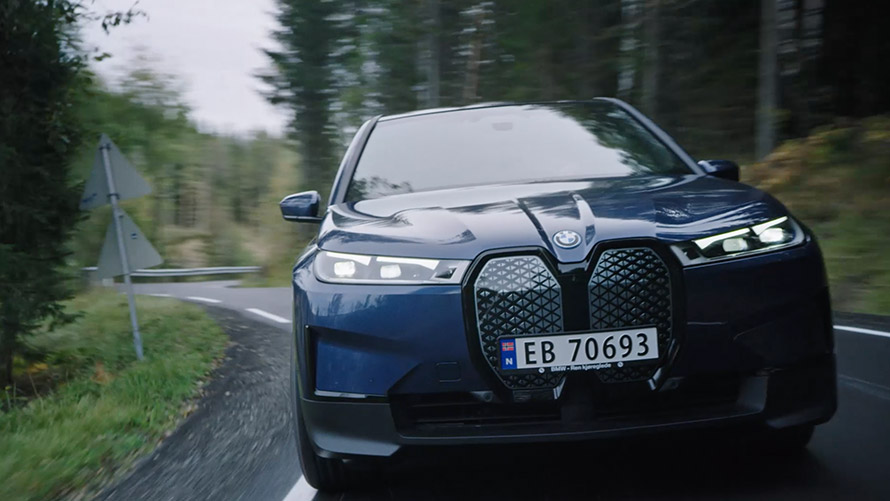 Žena v hnědém obleku se naklání proti BMW iX1 U11 2022 Žena v hnědém obleku se naklání proti BMW iX1 U11 2022 pohled zepředu vedle nabíjecí stanice