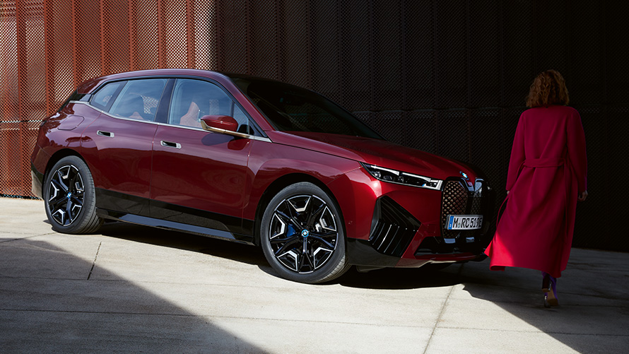 BMW Digitální klíč Plus BMW iX tříčtvrteční pohled zepředu před ženou BMW Digitální klíč Plus BMW iX tříčtvrteční pohled zepředu před ženou s černým kabátem