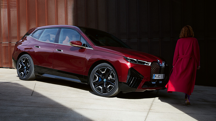 Žena před BMW iX i20 2021 tříčtvrteční pohled zepředu Žena v červeném kabátu před BMW iX i20 2021 tříčtvrteční pohled zepředu