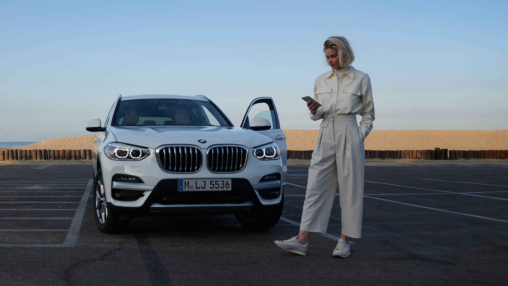 Žena používá aplikaci My BMW na chytrém telefonu. Žena používá aplikaci My BMW od BMW ConnectedDrive na chytrém telefonu před bílým BMW X5.
