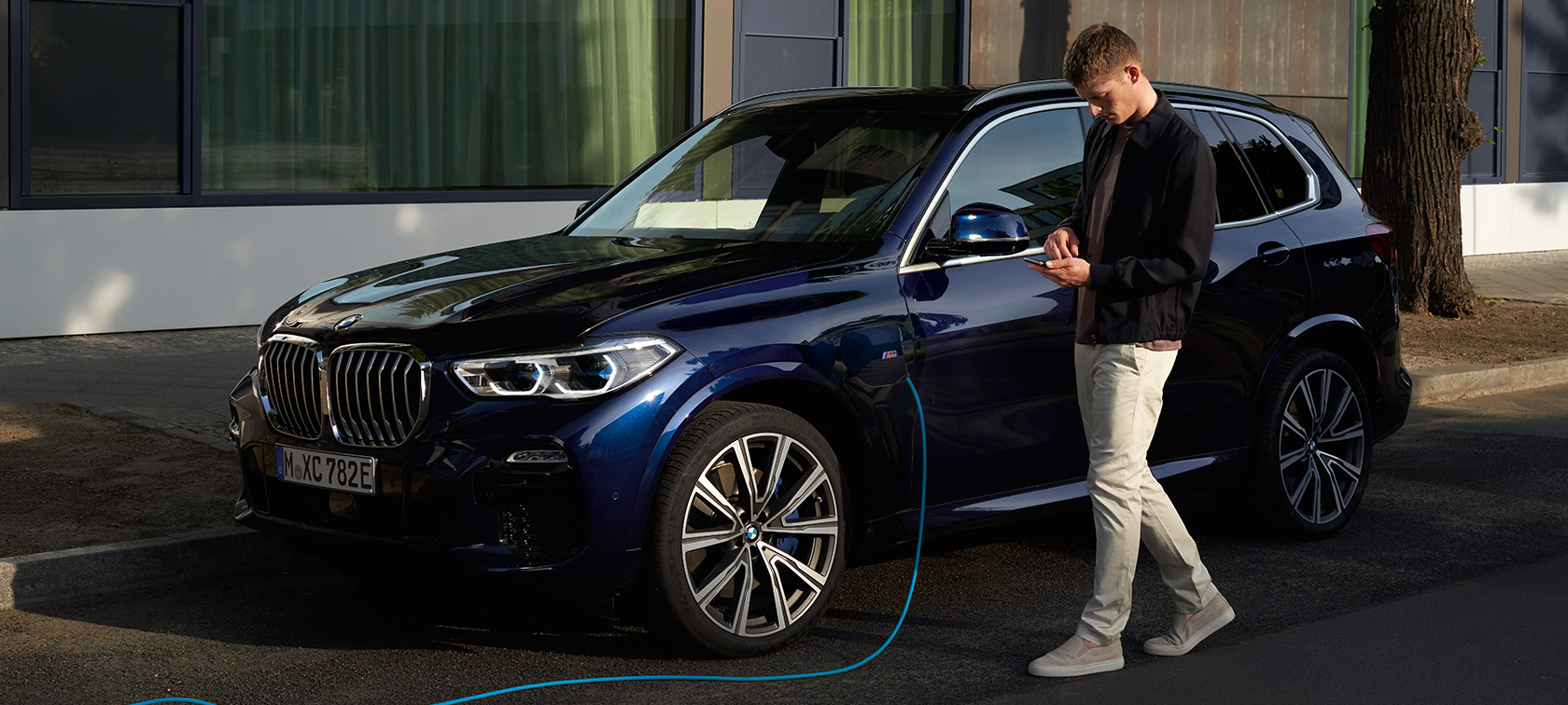 Muž používá aplikaci My BMW na chytrém telefonu. Muž používá aplikaci My BMW od BMW ConnectedDrive na chytrém telefonu při nabíjení jeho plug-in hybridního BMW X5
