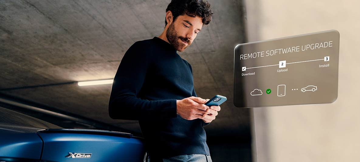 Muž opřený o záď BMW se dívá do svého telefonu Remote Software Upgrade Muž opřený o záď BMW se dívá do svého telefonu
