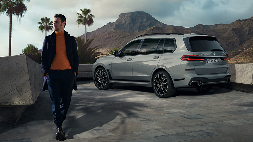 Muž stojící před zaparkovaným BMW X7 (G07) Dálkově ovládané parkování Dálkově ovládané parkování Muž stojící před zaparkovaným BMW X7 (G07) tříčtvrteční pohled zezadu pod palmou s horskou krajinou v pozadí