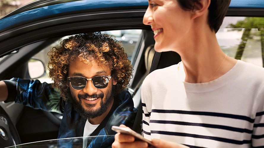 BMW Connected Muž v BMW a žena venku používají na svých chytrých telefonech BMW Connected.