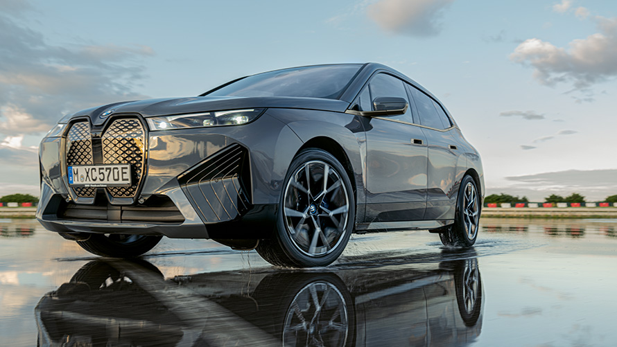 BMW Kola, Tříčtvrteční pohled zepředu na BMW iX (i20) na mokré silnici BMW Kola, Tříčtvrteční pohled zepředu na BMW iX (i20) na mokré silnici