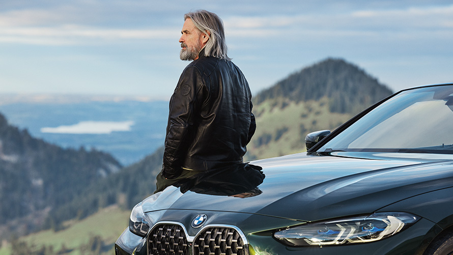Muž se sklání proti BMW před horskou scenérií BMW Příslušenství pro jaro a léto Muž se sklání proti BMW před horskou scenérií
