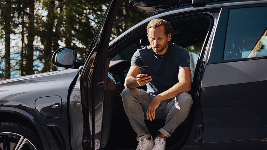 Muž sedí v BMW a dívá se do svého chytrého telefonu BMW Příslušenství pro jaro a léto Muž sedí v BMW a dívá se do svého chytrého telefonu