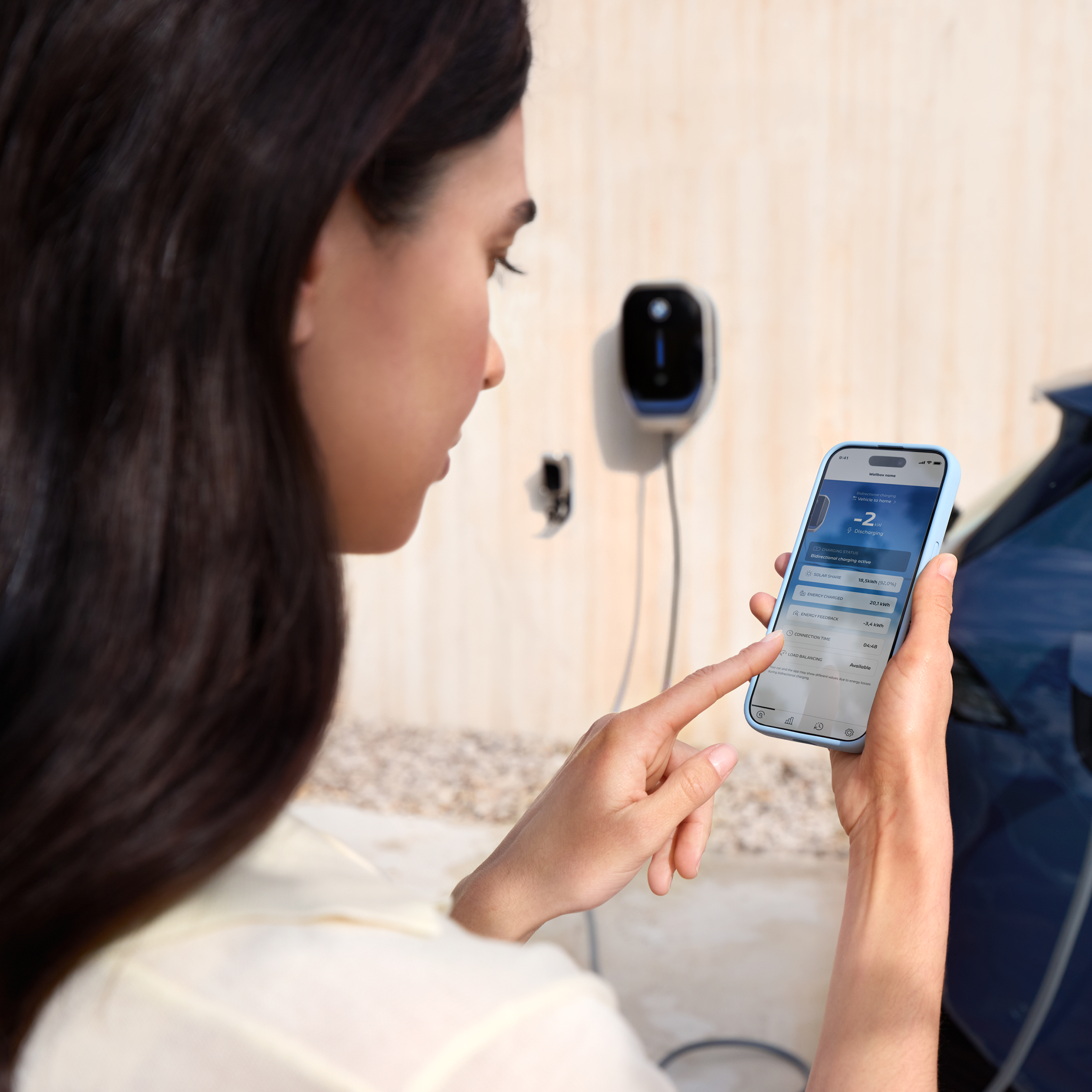 Žena se dívá na svůj chytrý telefon, na pozadí je vidět wallbox